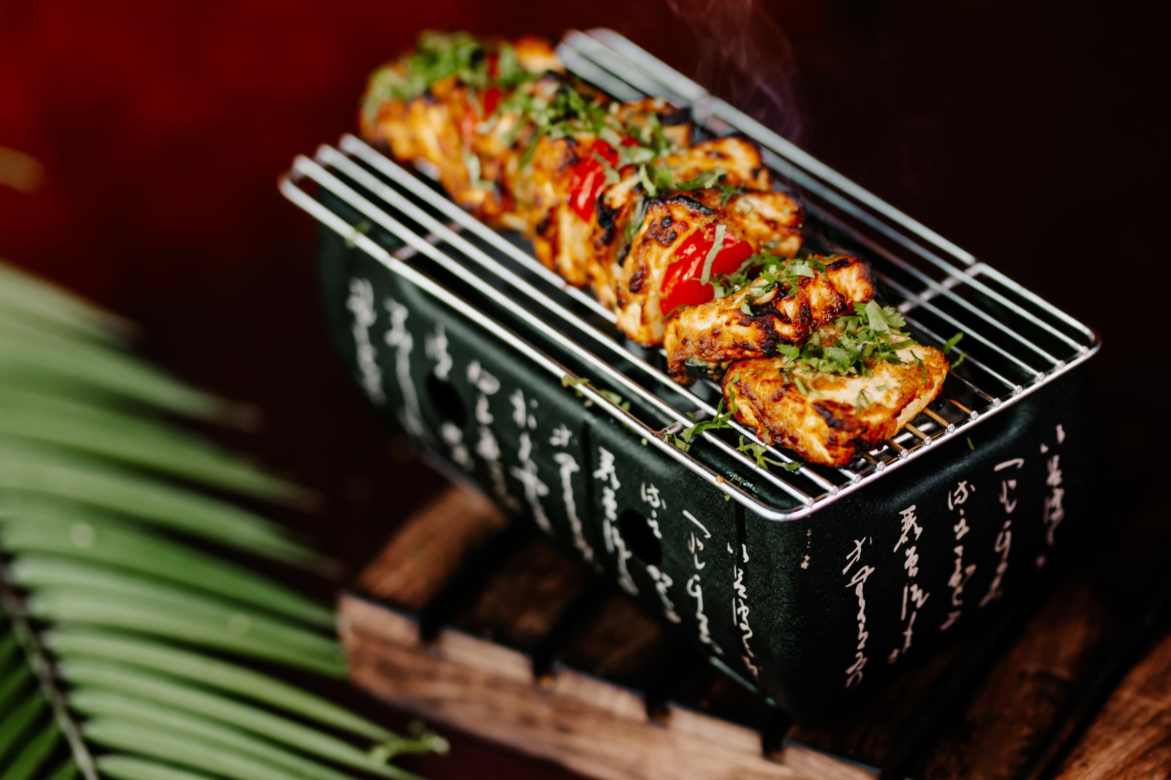 Grilled skewers on a black grill tray beside a palm leaf